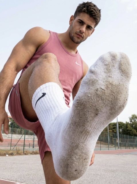 A muscular jock in pink sportswear extends his dirty white crew sock toward the camera in a POV perspective on an outdoor track.