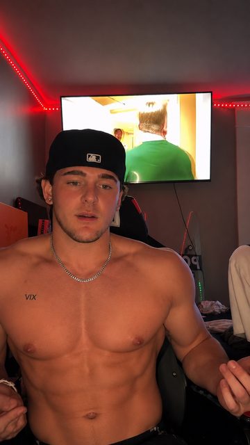 A shirtless muscle stud with a silver chain and a black baseball cap poses in a room with red ambient lighting.