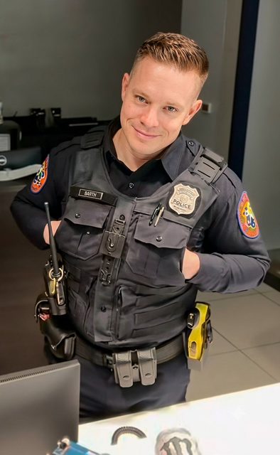 A man wearing a complete police uniform with a tactical vest and utility belt stands in an office setting.