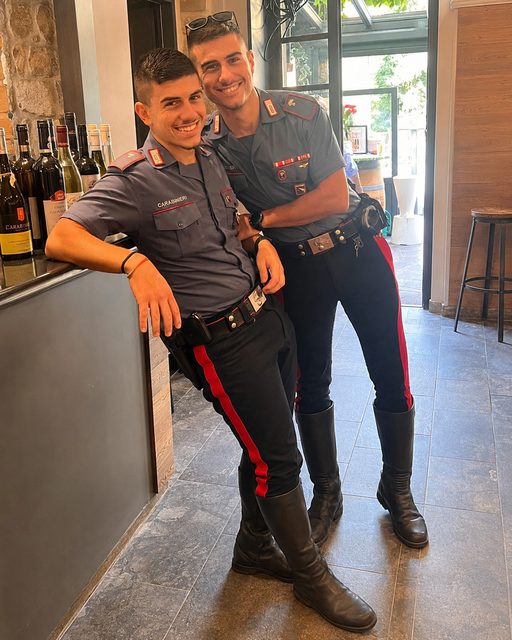 Two fit men in Carabinieri uniforms, featuring tall black leather boots and utility belts, pose together in an indoor setting.