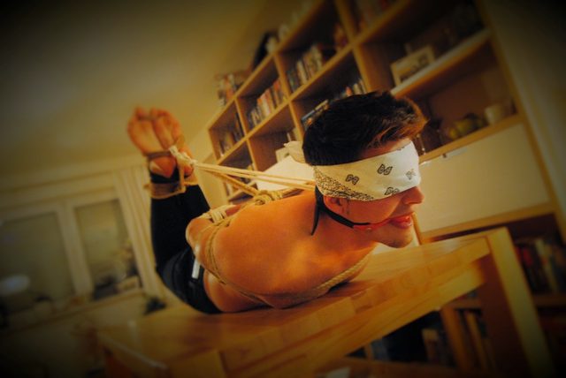 A shirtless young man lies prone on a wooden table, restrained in a rope hogtie with a blindfold and ball gag.