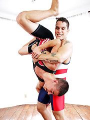 Two muscular men in wrestling singlets engage in a grappling session where one man lifts the other upside down.