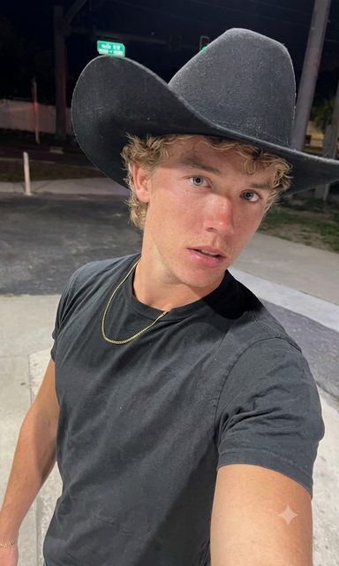 A young blonde twunk wearing a black cowboy hat and a black t-shirt takes a selfie outdoors at night.