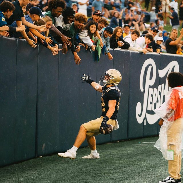 A muscular athlete in full football gear celebrates with fans at a stadium while holding a football.