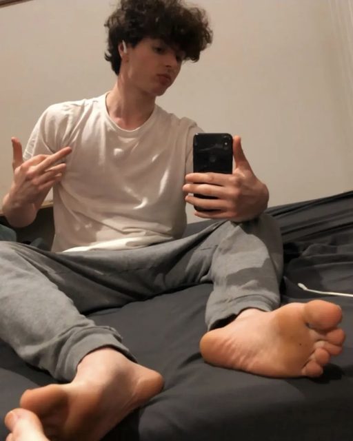 A young man takes a selfie in a bedroom, showing his bare feet.