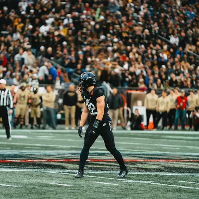 A muscular athlete wearing a full Navy football uniform and helmet stands on the field during a game with a large crowd in the background.