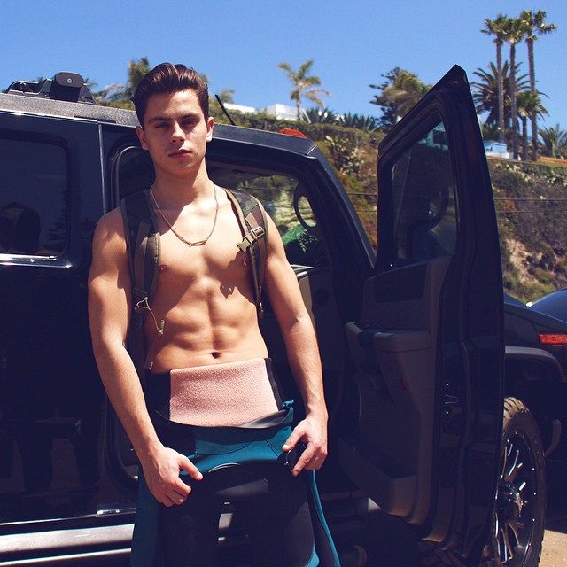 A muscular young man stands shirtless by a black car, wearing a wetsuit bottom and a backpack.
