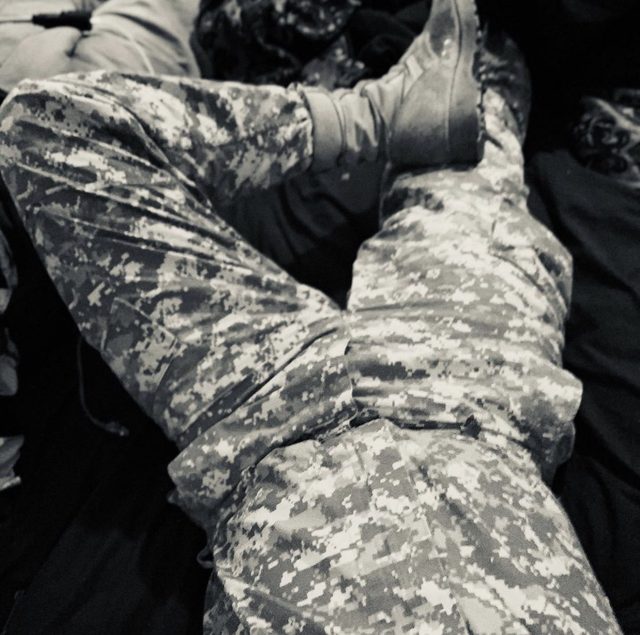 A man lies on a bed wearing digital camouflage trousers and combat boots.
