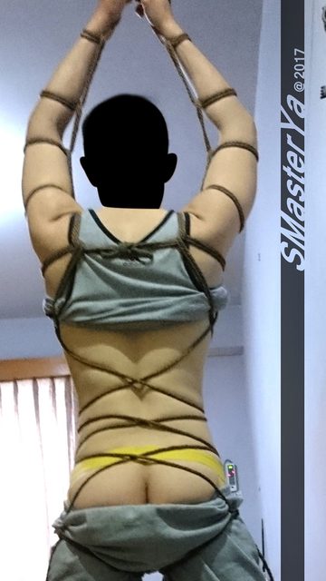 A person with raised bound arms and exposed lower back and buttocks bound in detailed shibari rope patterns indoors.