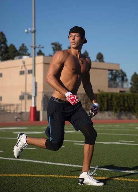 A shirtless twunk in gym shorts and football gloves runs across an outdoor turf field.