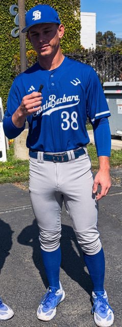 A muscular jock in a blue and gray Santa Barbara baseball uniform poses outdoors, showcasing a prominent bulge in his tight pants.