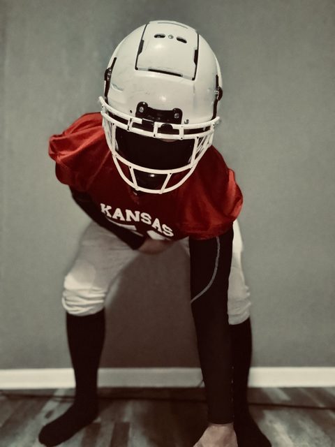 A man posing in a full Kansas football kit including a white helmet, red jersey, and compression sleeves.
