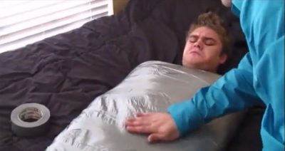 A young man lies on a bed wrapped tightly from the chest down in silver duct tape during a bondage session.