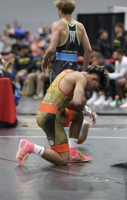 A young man wearing a patterned wrestling singlet and pink shoes kneels on a competition mat.