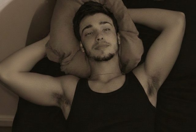 A young man with a trimmed beard and dark hair lies on a bed wearing a black ribbed tank top with his arms behind his head.