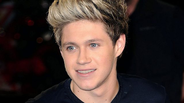 A close-up portrait of a smooth young man with styled blonde hair and blue eyes wearing braces and a dark shirt.
