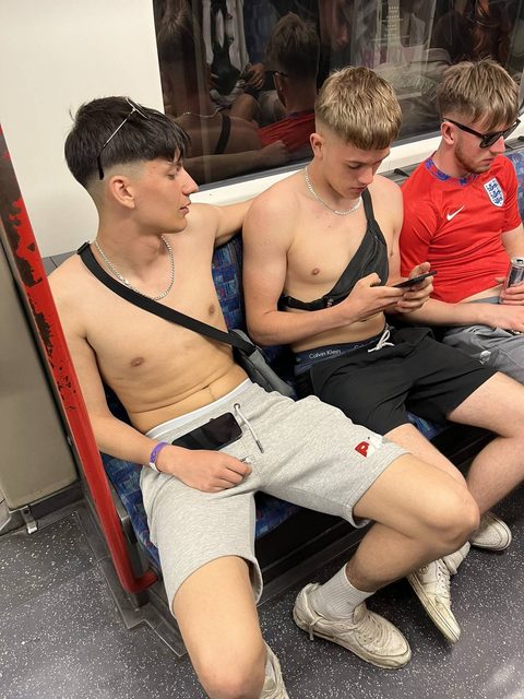 Three young shirtless men in shorts and sneakers sit together on a public train carriage, showing their underwear and silver chains.