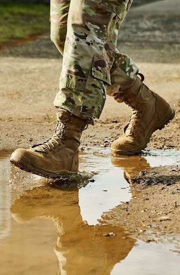 A person wearing camouflage military pants and tan combat boots walks through a muddy puddle outdoors.