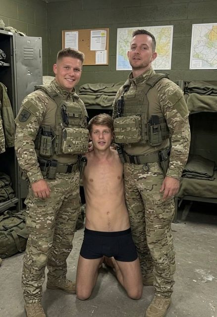 A shirtless man in boxer briefs kneels on the floor between two men dressed in full military camouflage uniforms and tactical gear inside a barracks.