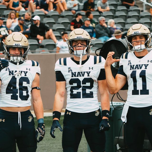 Three muscular football players in Navy uniforms and helmets stand on a stadium sideline.