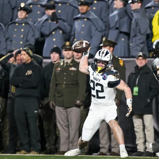 A muscular football player in a full Navy kit and helmet prepares to throw a ball in front of a military crowd.