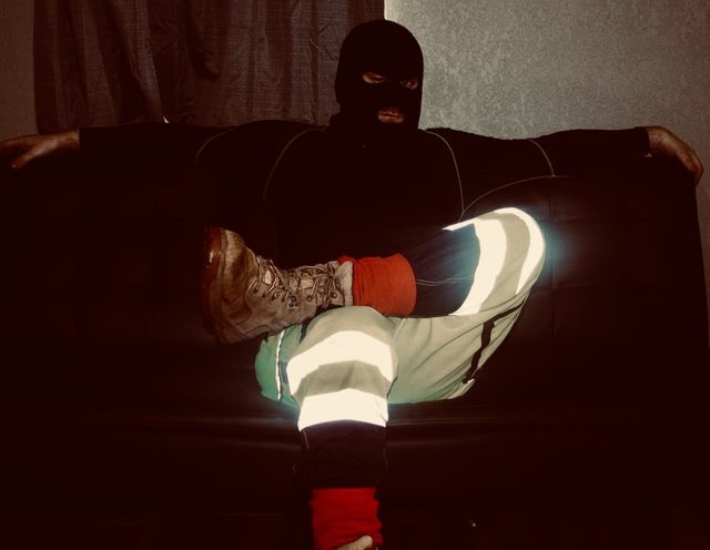 A man wearing a black balaclava, compression top, and reflective construction trousers sits on a dark couch in work boots.