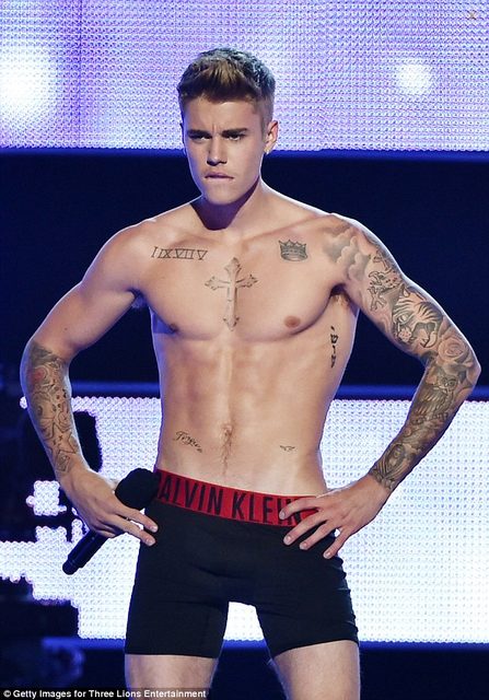 A shirtless man with tattoos performs on stage in black boxer briefs.