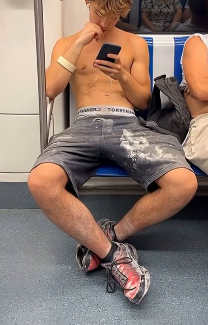 A shirtless young man with defined abs sits on a subway train using his smartphone while wearing paint-covered shorts.