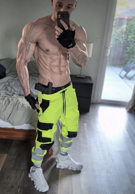 A shirtless, shredded muscle stud takes a mirror selfie in a bedroom wearing high-visibility cargo pants and fingerless gloves.