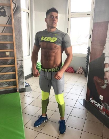 A muscular man poses in a gym setting wearing grey and yellow compression tights and a pulled-up shirt displaying his abs.