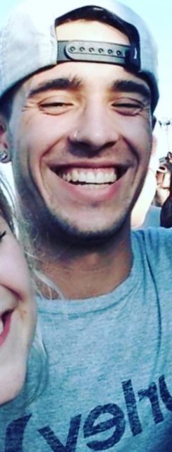 A smiling young twunk wearing a backwards snapback cap and a grey t-shirt at an outdoor event.
