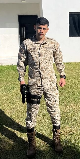A young man posing in a full desert camouflage tactical uniform with a thigh holster and combat boots outdoors.