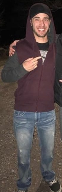 A smiling, bearded man in a maroon sleeveless hoodie and blue jeans stands outdoors at night.