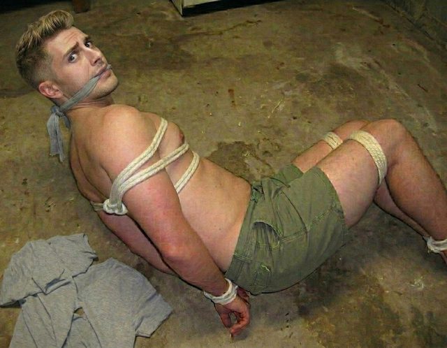 A muscular man is bound in rope and gagged with a cloth while lying on a concrete floor.