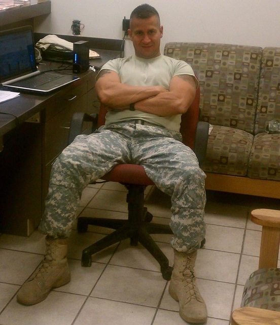 A muscular man in a military camouflage uniform and combat boots poses with arms crossed and legs spread wide in an office.
