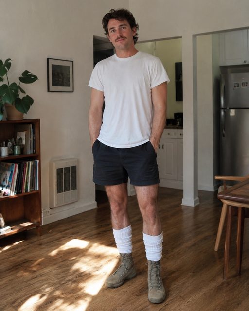 A fit man with a mustache poses in a home setting wearing gym shorts, tall white tube socks, and tactical combat boots.