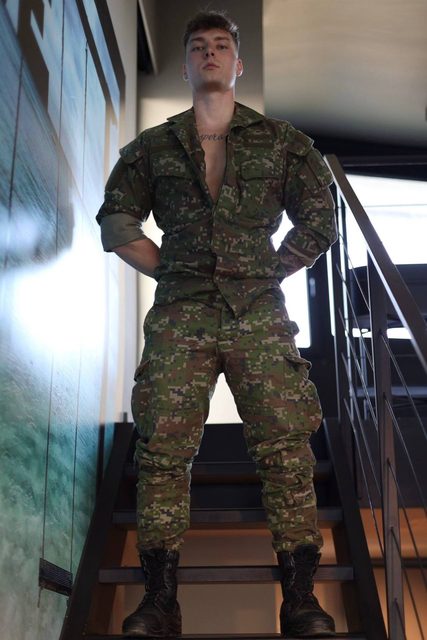 A muscular young man in a digital camouflage military uniform and black combat boots stands on a staircase with his hands behind his back.