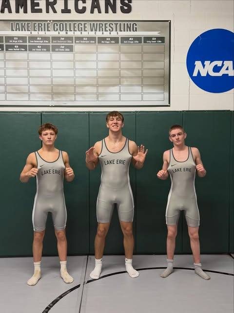 Three young men in matching grey college wrestling singlets pose together inside a wrestling room.
