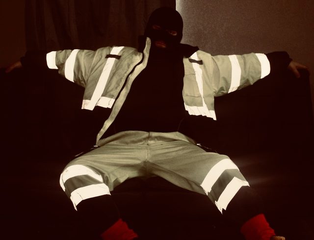 A person wearing high-visibility construction workwear and a black balaclava sits on a sofa in a dark room.