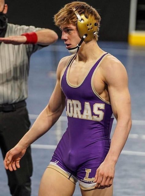 A young muscular wrestler wearing a tight purple spandex singlet and headgear stands on a gym mat.