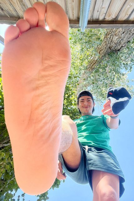 A young man outdoors holds his bare sole directly to the camera lens while dangling a blue sock.