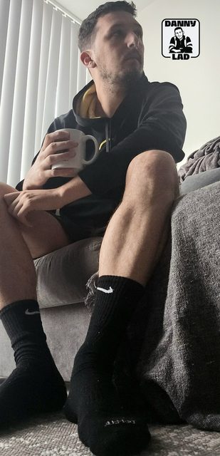 A bearded man in a black hoodie and shorts sits on a sofa holding a mug, showcasing his muscular legs and black Nike crew socks.