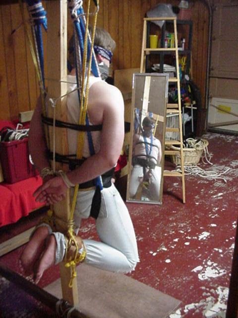 A man wearing denim jeans and a tank top is securely bound and suspended on a wooden post using ropes, straps, a blindfold, and duct tape gag.