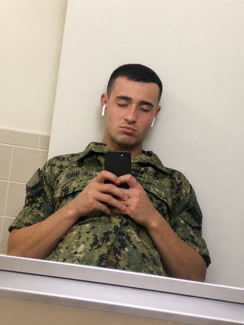 A young man with a buzzcut wearing a US Navy camouflage uniform takes a mirror selfie in a bathroom.