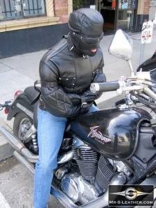 A person wearing a black leather straitjacket and a matching full leather hood sits bound atop a motorcycle on a city sidewalk.