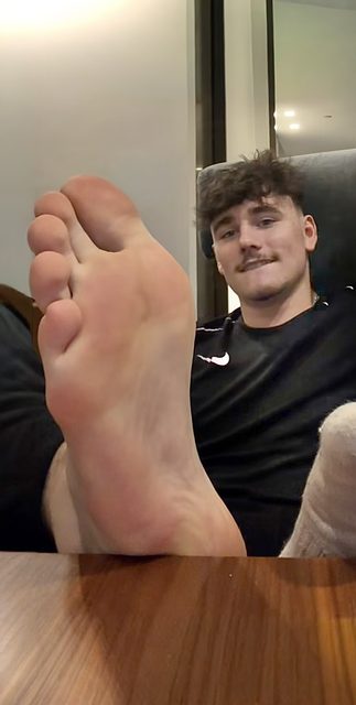 A young man with a mustache wearing a black athletic shirt presents his bare sole to the camera.