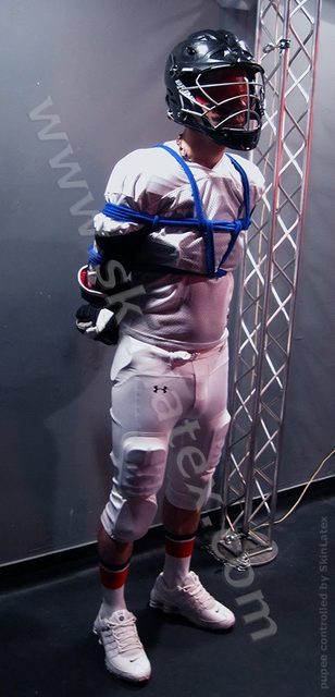 A man in white athletic gear, including a helmet and padded pants, stands bound in blue rope bondage next to a metal truss.