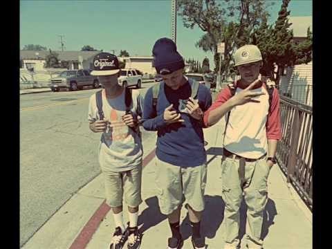 Three young men in casual streetwear, including baseball caps, beanies, and backpacks, pose on a sunny outdoor sidewalk.