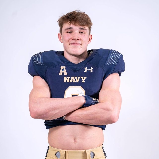 A muscular young man in a Navy football uniform with shoulder pads crosses his arms and exposes his midriff.
