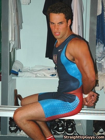 A muscular man in a tight red, blue, and white Nike wrestling singlet sits on a locker room bench with his hands behind his back.